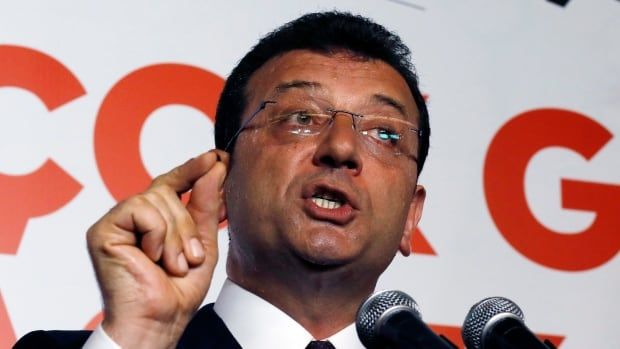 A man in a suit wearing glasses speaks in front of two microphones and gestures with his right hand.