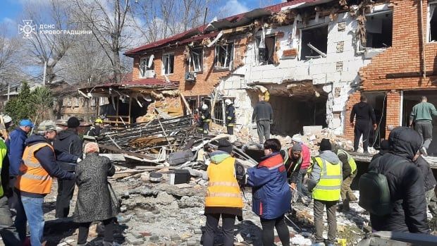 Several people are shown in the foreground, some in jackets and some in reflective safety vests, near a low-rise building significantly admaged, with concrete debris around it.