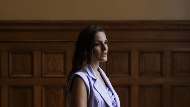 A woman in a lilac vets stands in front of a wodden panelled wall.