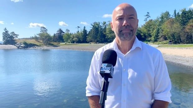 A white, balding man in a white dress shirt stands in front of a microphone with a stone beach, water and trees behind him