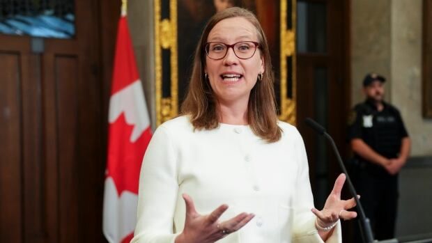 Karina Gould stands at a podium waring a white sweater as she speaks to members of the press in the Foyer of the House of Commons. 