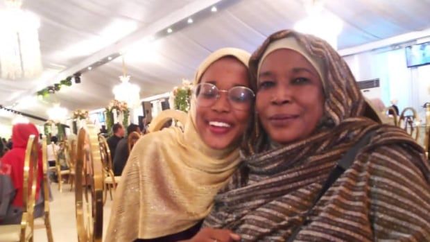 Two women in headscarves, one older and one younger, sit side by side in a restaurant, snuggling into each other and smiling 