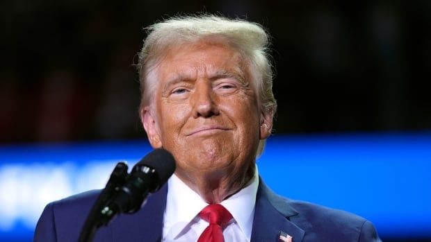 A man in a blue suit with a red tie smiles on stage at a political rally.