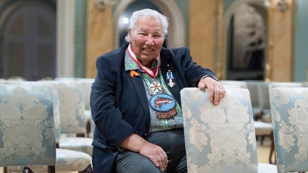 A man decorated in medals sits on a patterned chair while his left arm rests on the top of another chair in front of him. 