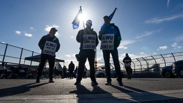 Three people wearing protest signs face the camera against the backdrop of a blue, sunny sky. The signs read "ILWU Local 514 Locked Out."