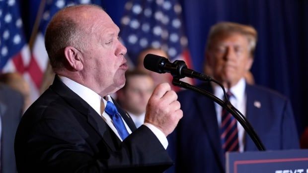 A man in a suit gestures while speaking at a lectern. Other men, and some American flags, are visible in the blurry background. 