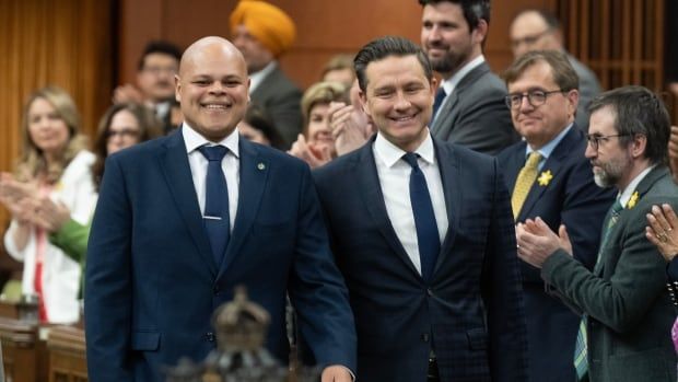 A bald man wearing a blue suit walks down the House of Commons with Pierre Poilievre