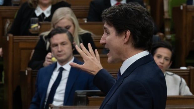 A man in a suit gestures with his hand as he speaks in the House of Commons. Another man looks on and listens.