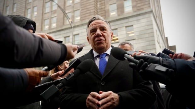 Man in black jacket and blue tie with lots of microphones pointed at him.