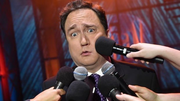 A man with dark hair and a suit has a confused look on his face as seven hands shove microphones into his face in a comedic way