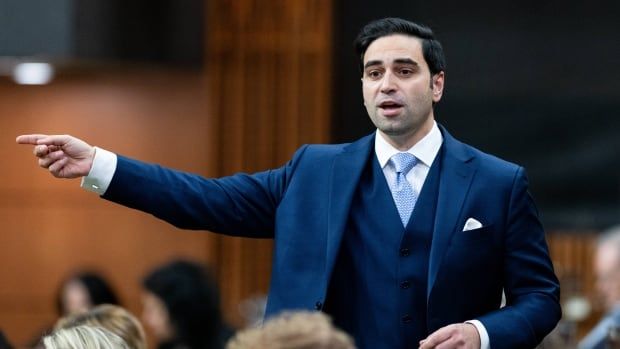 A man wearing a blue, three-piece suit points off camera in the House of Commons.