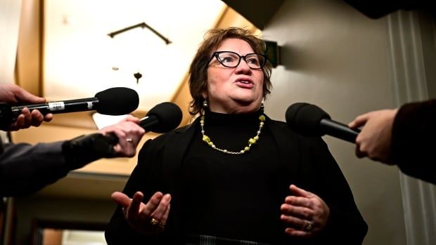 A woman wearing a black turtle neck and gold necklace speaks as reporters hold microphones.