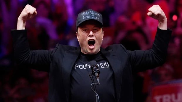 Elon Musk stands at a microphone on stage wearing a black suit jacket, black T-shirt and black Make America Great Again hat as he flexes both biceps and shouts to the crowd. 