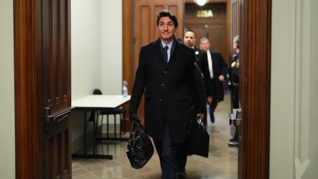 A man in a dark suit and black coat walks through doors inside a government building carrying a black briefcase.