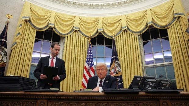 Trump at desk, surrounded by documents to sign and golden curtains.