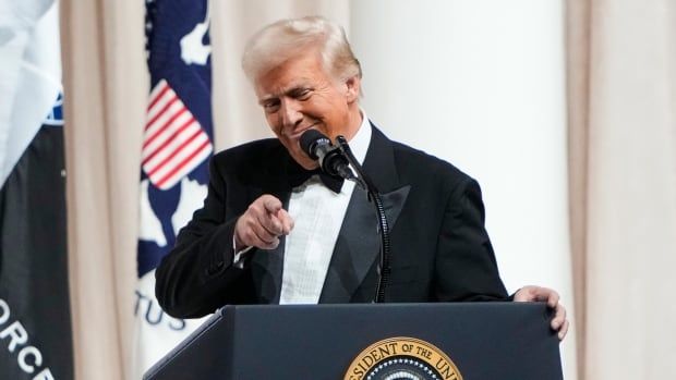 A man in a tuxedo, standing at a lectern, points and smiles at someone off camera.