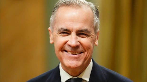 A man in a grey suit smiles in front of a yellow background.