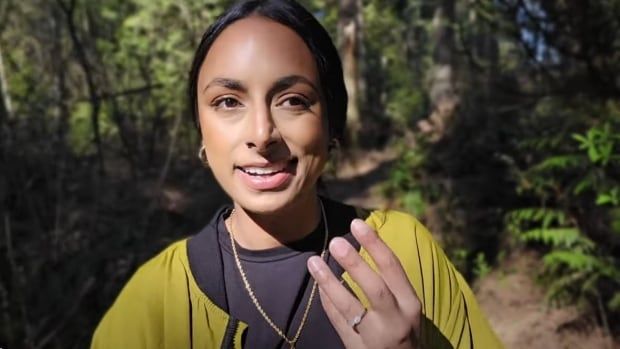 A young woman with long dark hair parted down the middle poses in a forest, wearing a yellow sweater over a dark top.