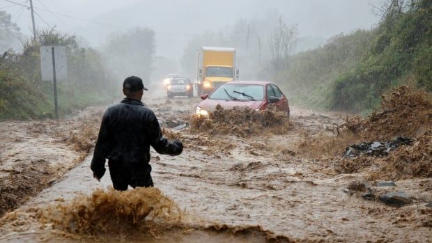 A man in a baseball cap with his back to the camera stands in muddy waters up his legs as motorists approach in a storm.