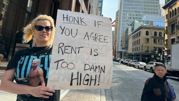 A blonde woman holds a sign that says 'Honk if you agree rent is too damn high!'