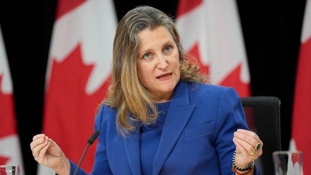 Freeland sits behind microphone in a blue suit. Canadian flags behind her