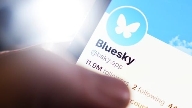 A closeup of a hand holding a phone with a Bluesky homepage on its screen.