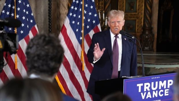 A man in a suit with a red tie speaks at a podium in front of American flags.