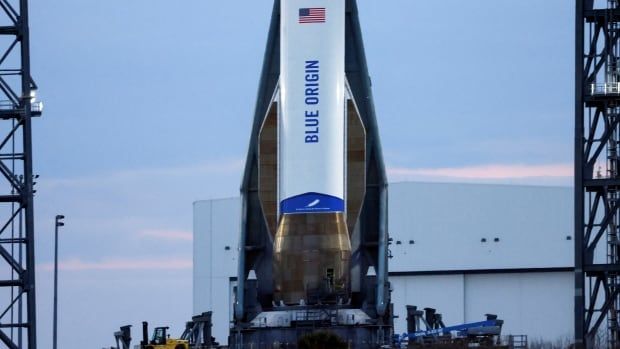 A white rocket, bearing a sign saying, "Blue Origin" sits on a launch pad.