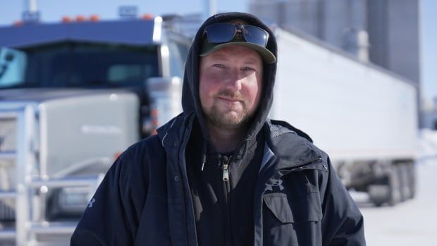 A farmer with a jacket and sunglasses looks into the camera with a semi-truck behind him.