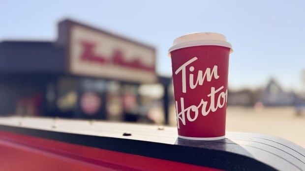 A Tim Hortons takeout cup sits on top of a Tim Hortons exit sign, with a Tim Hortons store visible in the background.