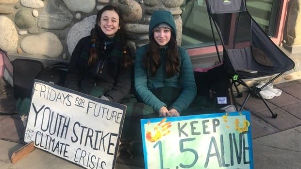 Two teenage girls sit on the ground holding homemade signs reading "Fridays for Future, Youth Strike for the Climate Crisis." 