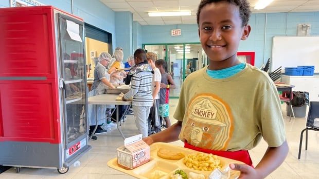 A smiling boy holds a lunch tray of milk, macaroni and cheese, steamed veggies, a piece of bread and cooked fruit, with other students serving and collecting lunch behind him. 