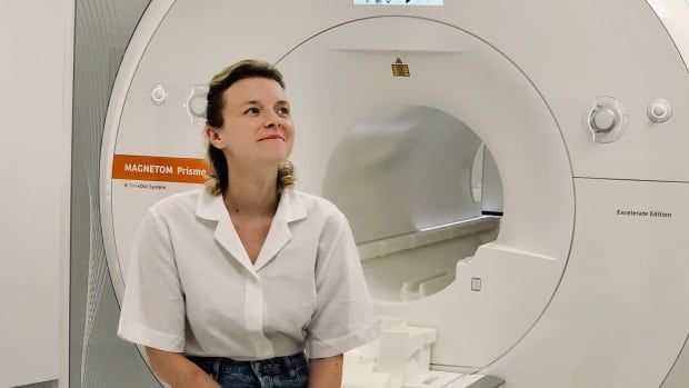 A woman in blue jeans in a white blouse sits on the edge of an MRI machine, her ankles crossed, staring off to one side and smiling. 