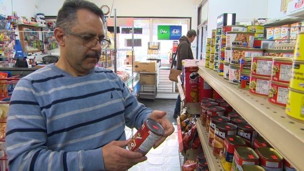 A shop owner looks at a can of food while standing in an aisle of his store. 