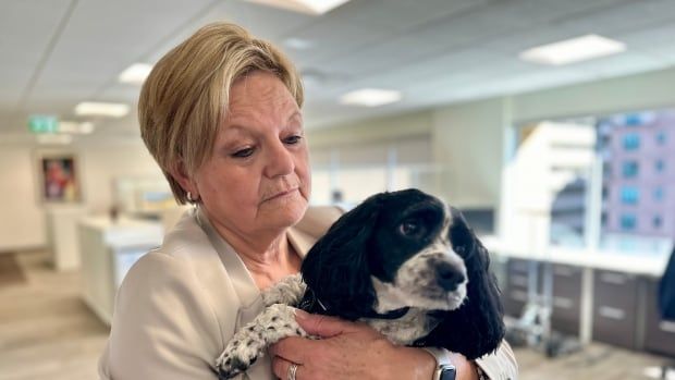 A woman with short, blonde hair wearing a beige blazer stands in an office holding a small black and white dog in her arms. 