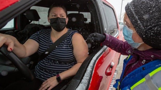 A doctor wearing wearing a winter coat and toque administers a vaccine to a woman wearing a T-shirt and sitting inside a truck. It's a snowy day.