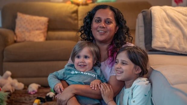 A mom holds her two children — 4-year-old Manny and 9-year-old Jasmine — in her arms. All three are sitting on the living room floor, smiling.