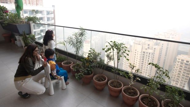 A woman crouches beside two children sitting on small chairs on a high-rise balcony. Beyond the balcony’s clear railing, heavy smog almost obliterates the city skyline. 