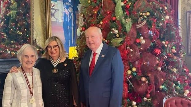 Three standing in front of a tall Christmas tree. one person has a medal around her neck.