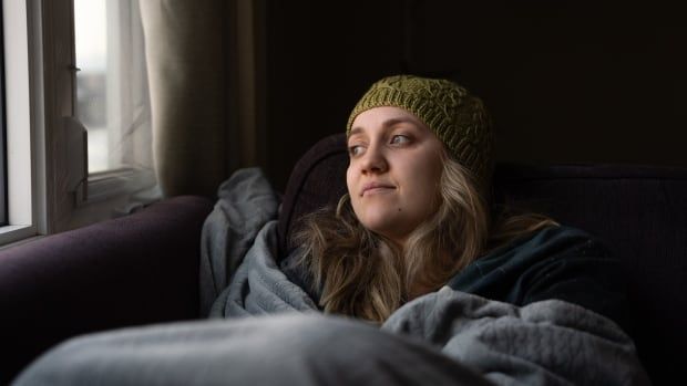 The photo shows a young woman, pale and tired, laying down on a couch covered with blankets and looking out the window. 