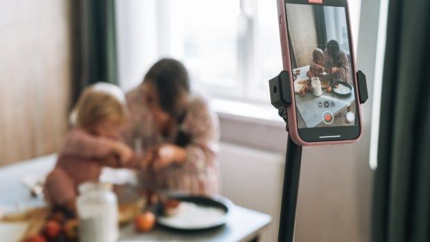 Young woman blogger cooking food with her baby daughter and recording video on mobile phone in the kitchen at home