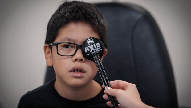 An optometry student holds a paddle to block the left eye of a boy wearing glasses, seated in an eye clinic. 