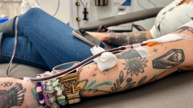 Blood is being drawn from a woman's arm, with bracelets, a watch and tattoos, with a machine in the background.