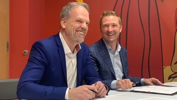 two men in suits sit at a table with pens