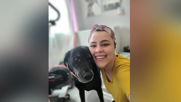 A woman wearing a wrap over her hair smiles widely in a photo with a black dog.