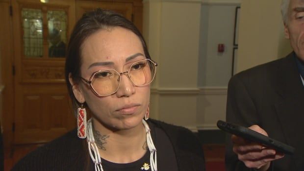 A woman with brown hair and glasses and beaded earring stands in the hallways of the legislature. 