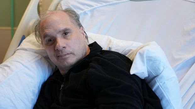 A man in a dark sweater sits in a hospital bed.