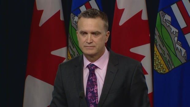 A man with grey hair stands in front of an Alberta flag at a podium.