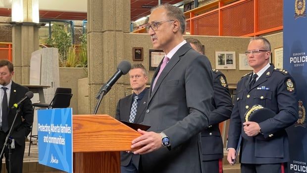 Mayor Amarjeet Sohi stands at a podium with a blue sign on the front of it. The sign reads: "Protecting Alberta families and communities." Sohi is wearing a dark grey or black suit with a light pink tie, as well as a smartwatch on his left hand. We are seeing him in profile.