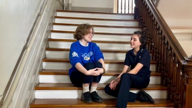 Two teenaged girls sit at the bottom of a set of stairs in a school hallway. They are wearing school uniforms and looking at each other as though in conversation.